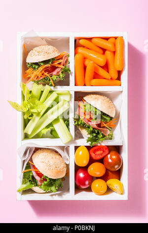 Hausgemachte kleine Burger mit Schinken, Tomaten, Möhren, frischem Salat in der alten weißen wooben Box auf rosa Hintergrund serviert. Gesunde junk food Konzept. Stockfoto