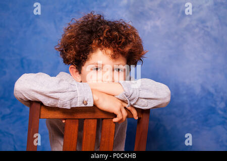 Portrait von cute Boy mit lockigem Haar nach hinten sitzen auf holzstuhl vor blauem Hintergrund und Kamera Stockfoto