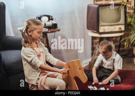 Süße kleine Mädchen auf Schaukelpferd und Bruder spielen mit Domino Fliesen hinter zu Hause sitzen, im Stil der 1950er Jahre Stockfoto