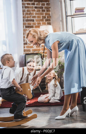 Glücklich, Mutter, Vater und Schwester an der niedlichen kleinen Jungen auf Schaukelpferd zu Hause sitzen suchen, 50s style Stockfoto