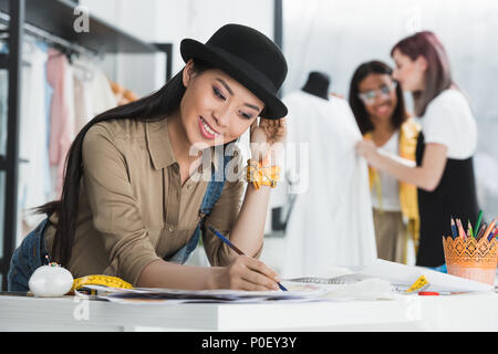 Junge lächelnde asiatische Modedesigner Skizzen während Kollegen hinter arbeiten Stockfoto