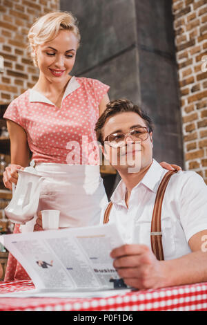 Lächelnde Frau in Schürze Kaffee zum Ehemann, lesen Zeitung am Tisch, im Stil der 1950er Jahre gießen Stockfoto