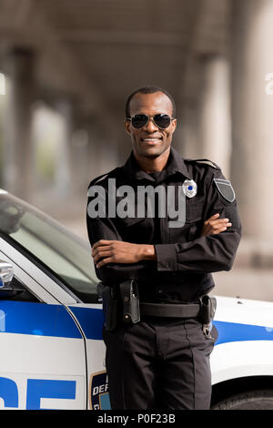Lächelnd afrikanische amerikanische Polizisten mit verschränkten Armen lehnte sich zurück auf Auto und Kamera Stockfoto