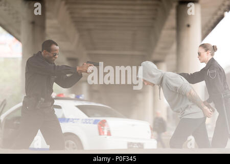 Seitenansicht der verärgerten Polizisten die Pistole auf die männliche Kriminelle, während ihn Partner Holding Stockfoto