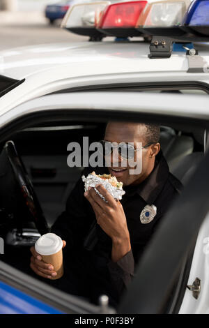 Junge afrikanische amerikanische Polizist mit Kaffee Tasse im Auto sitzen und Burger essen Stockfoto