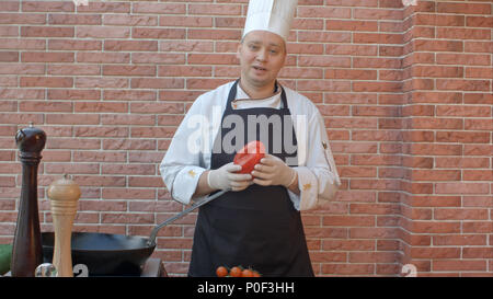 Im mittleren Alter Chef in weiße Uniform in der Nähe von Schreibtisch, im Gespräch mit der Kamera mit Gemüse in seine Hände, bereit, einen Salat vorbereiten Stockfoto