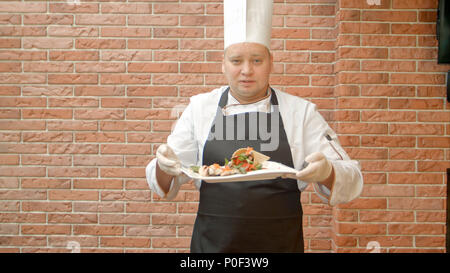 Freundliche Küchenchef in Uniform präsentieren eine Platte mit Meeresfrüchten Salat, im Gespräch mit einer Kamera Stockfoto