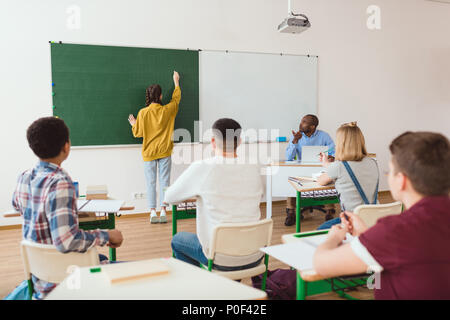 Ansicht der Rückseite des Schulmädchen schreiben auf kreidetafel und Klassenkameraden mit dem Lehrer im Klassenzimmer sitzen Stockfoto