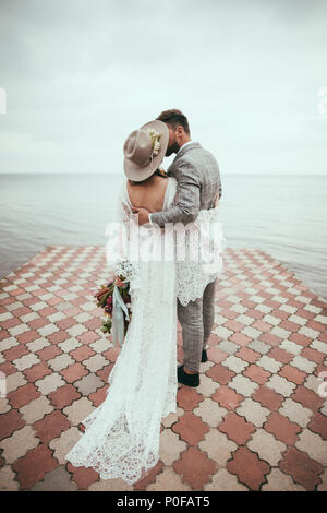 Rückansicht der Braut und Bräutigam in Boho style Umarmen und Küssen auf Pier am See Stockfoto