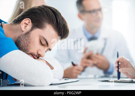 Erschöpft junger Arzt schlafen auf Schreibtisch, während Kollegen in Gespräch Stockfoto