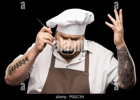 Professioneller Koch in Hut und Schürze Verkostung Gericht aus Schöpfkelle isoliert auf Schwarz Stockfoto