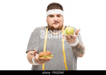Junge mollig Mann an der Kamera schaut, während Sie Burger und Apple auf Weiß isoliert Stockfoto