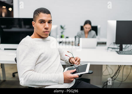 Hübscher junger Geschäftsmann mit Tablette zu Open Space Office sitzen Stockfoto