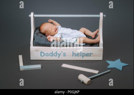 Kleines Baby in bodysuit Schreien während in Holz- Toolkit liegen mit Daddys helper Schriftzug Stockfoto