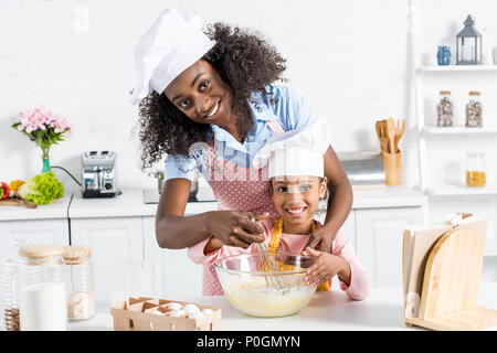 Afrikanische amerikanische Mutter und Tochter in Kochmützen mischen Teig mit Schneebesen auf Küche Stockfoto