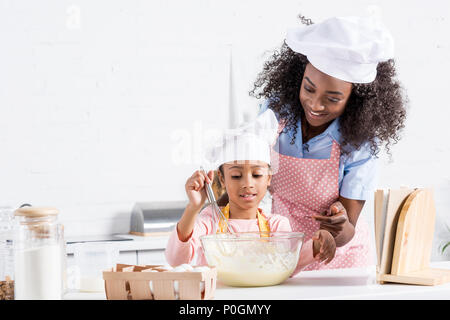 Afrikanische amerikanische Mutter und Tochter in Kochmützen mischen Teig auf Küche mit Kochbuch Stockfoto