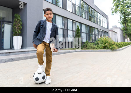 Cute african american scoolboy stehend mit Fußball, und wenn man die Kamera in der Nähe der Schule Stockfoto