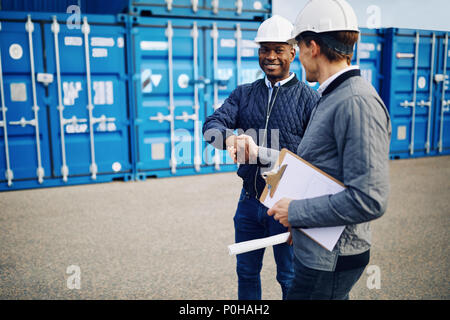 Zwei zusammen lächeln Ingenieure tragen hardhats Hände schütteln bei der Stellung von Containern auf einem kommerziellen Dock Stockfoto