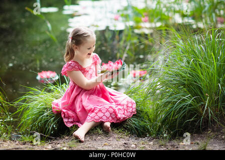 Kind am Seeufer sitzen beobachten Seerose Blüten. Kleines Mädchen mit rosa Lilie Blume. Zicklein an Lilien auf sonniger Frühlingstag. Kinder outdoor Stockfoto