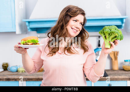 Portrait von übergewichtigen Frau mit Burger und frischem Brokkoli in Händen in der Küche zu Hause, gesunde Ernährung Konzept Stockfoto