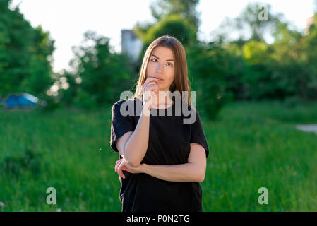 Brunette Mädchen im Sommer in einem Park im Freien. Er berührt seine Lippen mit einer Hand. Geistig träumt von der Zukunft. Emotionen einer Fantasie des Begehrens. In lässig gekleidet. Stockfoto