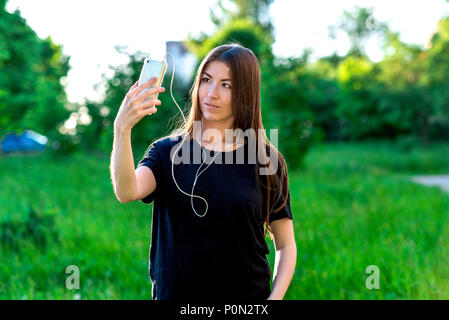 Asiatische Mädchen im Sommer im Park. Schöne Brünette. Lächelnd und mit Blick auf das Mobiltelefon Musik hören. Sendet die Sprachnachricht. Nehmen Sie Fotos auf dem Telefon. Stockfoto