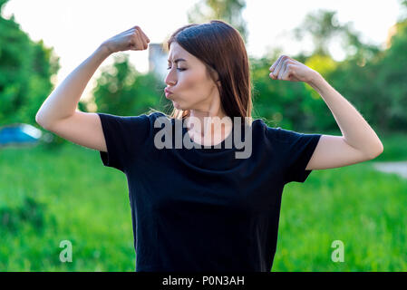 Die brünette Mädchen im Sommer in einem Park auf der Natur. Gesten der Hände Muskeln zu zeigen. Emotional er küsst. Zum Narren macht sich über die Stärke der Männer. In einem schwarzen T-Shirt. Stockfoto