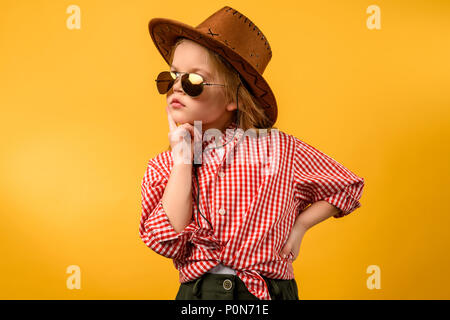 Stilvolle cowgirl in Hut und Sonnenbrille posiert, isoliert auf Gelb Stockfoto