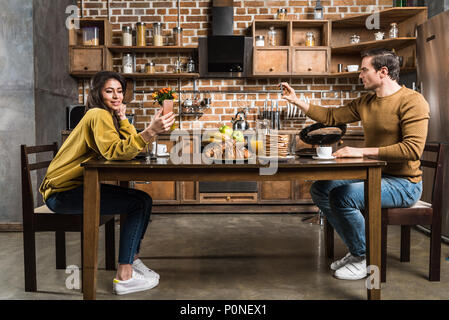 Seitenansicht des multiethnischen Paar mit Smartphones beim Frühstück Stockfoto