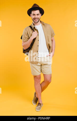 Die volle Länge der gutaussehenden jungen Mann in kurzen Hosen und Hut, Rucksack und lächelnd an der Kamera auf gelb isoliert Stockfoto