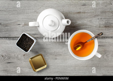 Table Top - Messing Tee Caddy, weißes Porzellan Teekanne und Tasse aus Keramik mit silbernen Löffel und heiße Amber trinken, auf grau Holzschreibtisch. Stockfoto
