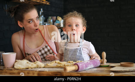 Junge Mutter und Tochter bereiten Sie Cookies in der Küche. Sie sind in den Schürzen. Kleines Mädchen und Mutter Überraschung und Bewunderung. Positive Emotionen. Familie ti Stockfoto