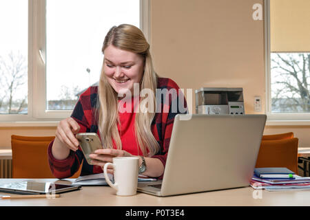 Eine lächelnde blonde Frau an einem Laptop sitzt und spricht über ein Handy. Stockfoto