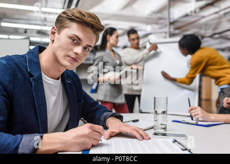 Hübscher junger Geschäftsmann unterzeichnen Vertrag während im Hintergrund arbeitenden Kollegen Stockfoto