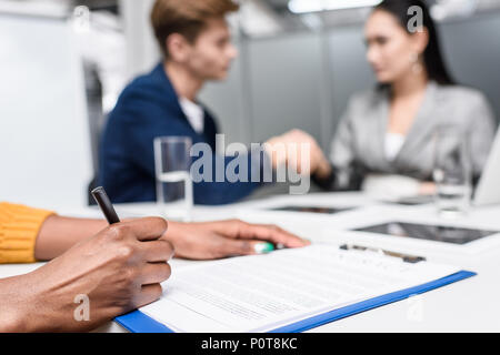 7/8 Schuß von afrikanische amerikanische Frau unterzeichnen Vertrag während verschwommene Geschäftspartner Hände schütteln auf Hintergrund Stockfoto