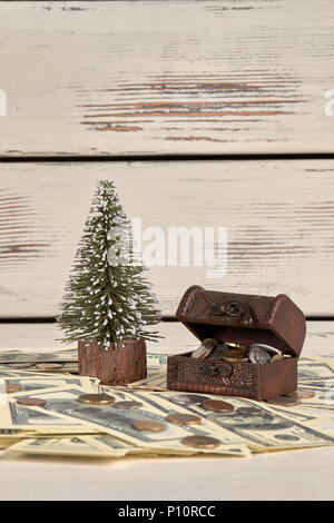 Haufen Geld, kässeli Brust und Weihnachtsbaum. Dollar und Münzen. Weiß Holz Hintergrund. Stockfoto