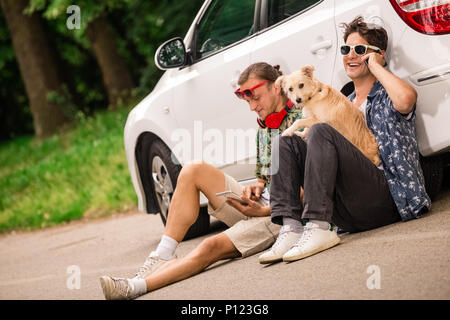Hipster Jungs und ihr Hund sitzt neben dem Auto auf der Straße und mit Handy und Tablet Gerät. Stockfoto