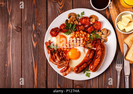 Traditionelles Englisches Frühstück mit Spiegelei, Würstchen, Bohnen, Pilze, gegrillten Tomaten und Speck auf Holz- Hintergrund. Ansicht von oben Stockfoto