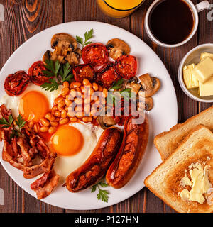 Traditionelles Englisches Frühstück mit Spiegelei, Würstchen, Bohnen, Pilze, gegrillten Tomaten und Speck auf Holz- Hintergrund. Ansicht von oben Stockfoto