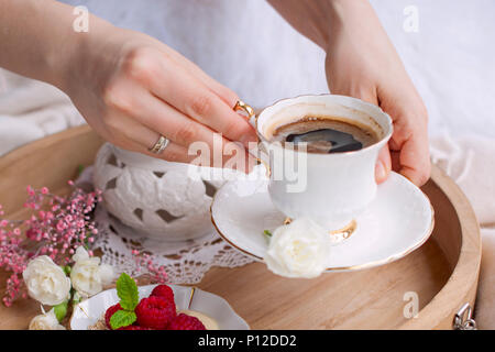 Eine Frau trinkt Kaffee im Bett. Eine hölzerne Fach mit Frühstück. Himbeere Beeren und Blumen. Helle Farben. Romantik. Kaffeetasse in den Händen. Stockfoto