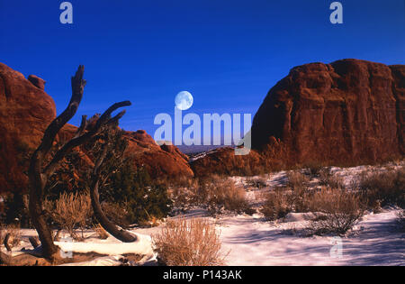 Arches National Park: Blick auf den fernen Horizont durch Felsformationen mit Vollmond über Schnee bedeckte Wüste im späten Licht, in der Nähe von Moab, Utah, USA Stockfoto