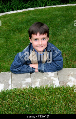 Eine nette Braunhaarige Kind lehnt sich gegen eine Mauer aus Stein im Gras und grinst Stockfoto