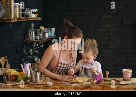 Junge Mutter und Tochter bereiten Sie Cookies in der Küche. Sie sind in den Schürzen. Kleine Mädchen und Frauen drücken Backformen. Zeit mit der Familie. Stockfoto