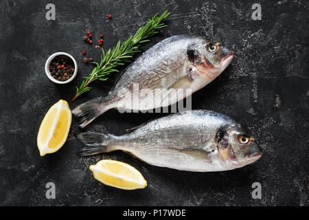 Frische dorado oder wolfsbarsch Fisch auf schiefer Hintergrund mit Zutaten zum Kochen, Zitrone, Rosmarin und Gewürzen. Ansicht von oben Stockfoto