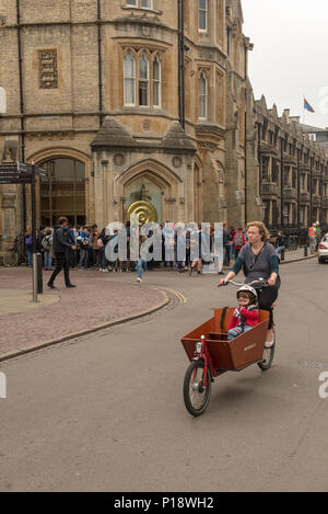 Frau fährt mit einem Kind auf einem Lastenrad an der Corpus Clock in Cambridge vorbei, während Touristen sich um die berühmte kinetische Skulptur und das Wahrzeichen versammeln Stockfoto