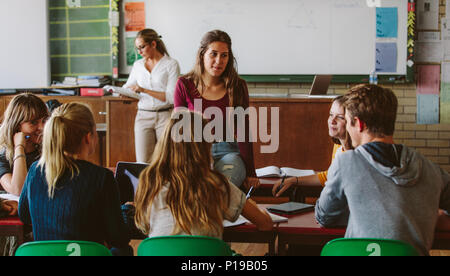 Junge Frau sitzt auf der Klasse Tabelle mit Mitschülern um und sprechen. Gruppe von Schülern der Auseinandersetzung mit neuen Aufgaben im Klassenzimmer. Stockfoto