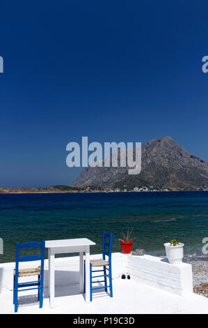 Tisch und Stühle mit Insel Telendos, in der Ferne, Myrties Kalymnos, Dodekanes, Griechenland. Stockfoto