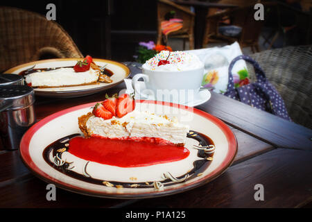 Käsekuchen mit frischen Erdbeeren auf Platte und einer Tasse Wiener Kaffee mit Schlagsahne auf braunem Holz- Tabelle in einen Außenpool im Sommer Cafe. Entspannen gemütliche Altstadtatmosphäre. offee brechen, Dessert. Stockfoto