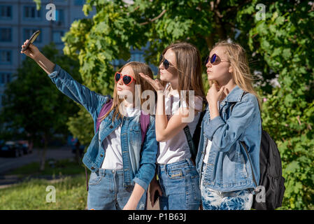 Drei Mädchen im Teenageralter im Sommer im Freien. Macht ein Foto von der Person am Telefon. Sie tragen schicke Jeans Kleidung. Sonnige Schutzbrille. Jugendmädchen sind Blondinen mit Rucksäcken hinter ihren Rücken. Stockfoto