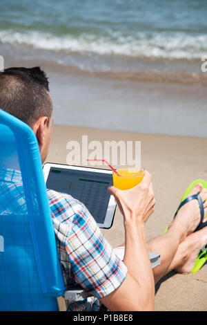 Nahaufnahme eines jungen kaukasischen Mann am Strand, mit einem digitalen Tablet, sitzend auf einem blauen Liegestuhl mit einem Glas einen erfrischenden Drink orange in seinem Stockfoto